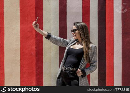 Content female in trendy outfit taking self portrait on mobile phone in street against colorful striped background. Stylish woman taking selfie in city on sunny day