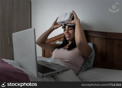 Content female putting on glasses of virtual reality while sitting on bed with laptop and preparing for experiencing cyberspace at home. Woman using VR goggles and laptop on bed