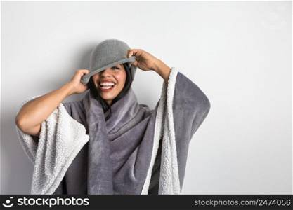 Content Hispanic female model wrapped in soft blanket laughing happily while covering eye with beanie against white background. Cheerful ethnic woman in blanket taking on gray hat