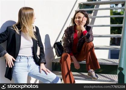 Content young girls best friends in stylish outfits enjoying sunny day standing on staircase in city park. Delighted young women smiling while resting near stairs in park