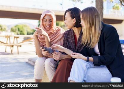 Content young multiracial female best friends, in casual clothes and hijab, smiling while watching video on smartphone sitting on bench in city park on sunny day. Positive multiethnic girlfriends sharing smartphone while resting on bench in park