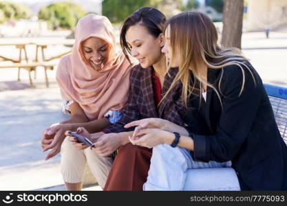 Content young multiracial female best friends, in casual clothes and hijab, smiling while watching video on smartphone sitting on bench in city park on sunny day. Positive multiethnic girlfriends sharing smartphone while resting on bench in park