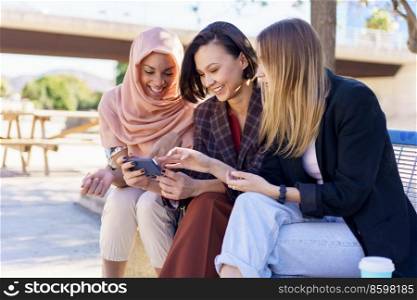Content young multiracial women best friends, in casual clothes and hijab, smiling while watching video on smartphone sitting on bench in city park on sunny day. Positive multiethnic women sharing smartphone while resting on bench in park