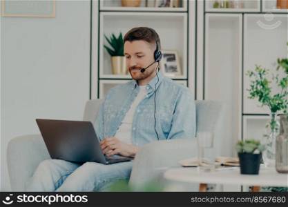 Contented guy in comfortable clothes using headset for video communication and holding laptop on his lap while staying home. Male freelancer working online. Distant work and freelance concept. Happy young man sitting and typing on laptop