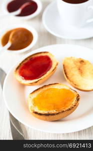 Continental breakfast consisting of toasted bread rolls with butter, peach and strawberry jam with tea in the back, photographed with natural light (Selective Focus, Focus in the middle of the first bread roll). Continental Breakfast