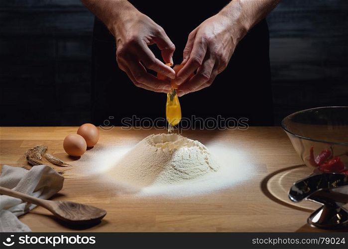 Cook hands kneading dough, cracking an egg in flour. Low key shot, close up on hands, some ingredients around on table.