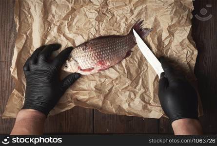 cook in black gloves cleans whole fish carp from scales on a brown piece of paper, top view