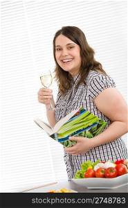 Cook - plus size happy woman holding cookbook and glass of white wine in kitchen