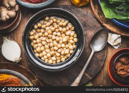 Cooked chick peas in dish with spoon and cooking ingredients on rustic wooden kitchen table background, top view. Healthy vegetarian or vegan food and eating concept