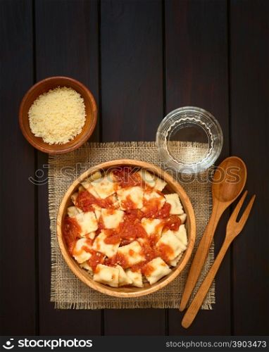 Cooked ravioli with homemade tomato sauce in wooden bowl with grated cheese and glass of water, wooden spoon and fork on the side, photographed overhead on dark wood with natural light