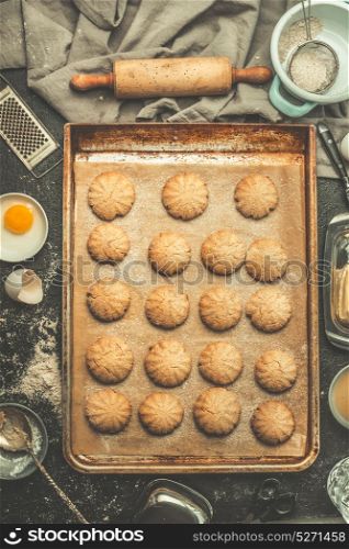Cookies on baking tray with rolling pin and dough ingredients on kitchen tables background, top view