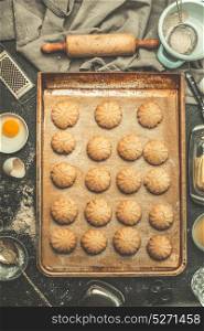 Cookies on baking tray with rolling pin and dough ingredients on kitchen tables background, top view