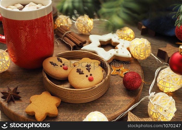 Cookies with a deer face in a wooden cup, red mug of coffee with marshmallows, gingerbread, garland, cinnamon, Christmas ball and fir branches on a wooden background.
