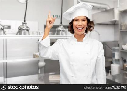 cooking, advertisement and culinary concept - happy smiling female chef in toque pointing finger up over restaurant kitchen background. female chef pointing finger up over restaurant