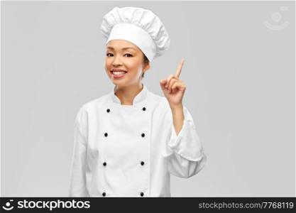 cooking, advertisement and food concept - happy smiling female chef in jacket pointing finger up over grey background. smiling female chef in jacket pointing finger up