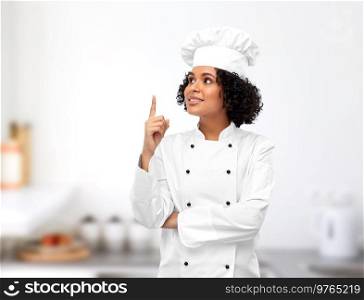 cooking, advertisement and food concept - happy smiling female chef in toque pointing finger up over restaurant kitchen background. smiling female chef pointing finger up in kitchen