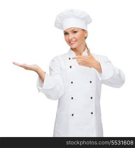 cooking, advertisement and food concept - smiling female chef, cook or baker holding something on palm of hand and pointing finger to it