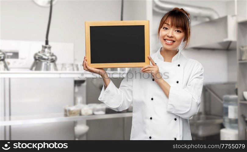 cooking, advertisement and people concept - happy smiling female chef holding black chalkboard over restaurant kitchen background. female chef holding black chalkboard on kitchen
