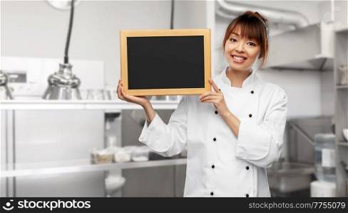 cooking, advertisement and people concept - happy smiling female chef holding black chalkboard over restaurant kitchen background. female chef holding black chalkboard on kitchen