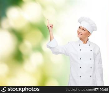 cooking, advertisement and people concept - smiling female chef, cook or baker pointing finger up to something over green background