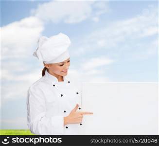 cooking, advertisement and people concept - smiling female chef, cook or baker pointing finger to white blank board over blue sky and grass background