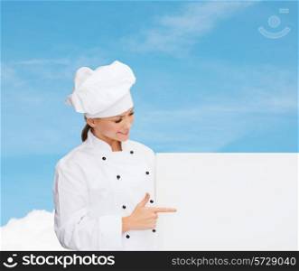 cooking, advertisement and people concept - smiling female chef, cook or baker pointing finger to white blank board over blue sky with cloud background