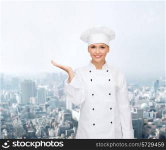 cooking, advertisement and people concept - smiling female chef, cook or baker holding something on palm of hand over city background