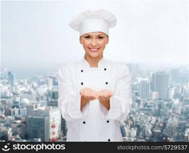 cooking, advertisement and people concept - smiling female chef, cook or baker holding something on palms of hands over city background