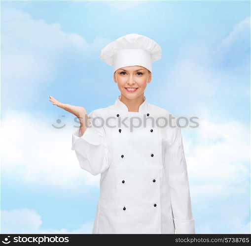 cooking, advertisement and people concept - smiling female chef, cook or baker holding something on palm of hand over blue cloudy sky background