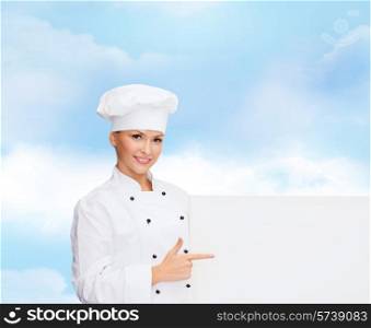 cooking, advertisement and people concept - smiling female chef, cook or baker pointing finger to white blank board over blue cloudy sky background