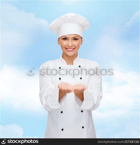 cooking, advertisement and people concept - smiling female chef, cook or baker holding something on palms of hand over blue cloudy sky background