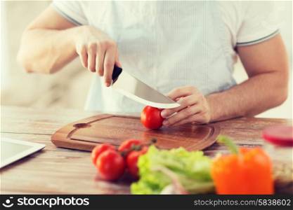 cooking and home concept - close up of male hand cutting tomato on cutting board with sharp knife