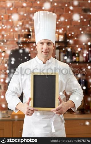 cooking and people concept - happy male chef cook with black blank menu chalk board in kitchen in restaurant kitchen over snow effect. happy male chef with blank menu board in kitchen