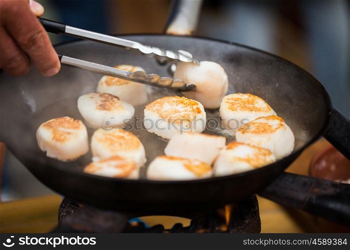cooking, asian kitchen, sale and sea food concept - close up of cook with tongs frying scallops in cast iron pan at asian street market