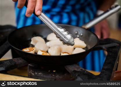 cooking, asian kitchen, sale and sea food concept - close up of cook with tongs frying scallops in cast iron pan at asian street market