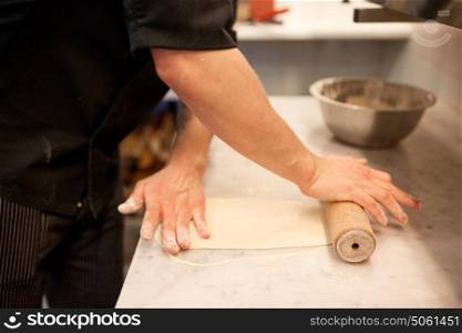 cooking, baking and people concept - close up of male chef with rolling-pin rolling dough at restaurant or bakery kitchen. chef with rolling-pin rolling dough at kitchen