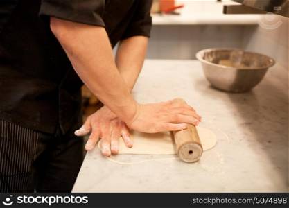 cooking, baking and people concept - close up of male chef with rolling-pin rolling dough at restaurant or bakery kitchen. chef with rolling-pin rolling dough at kitchen