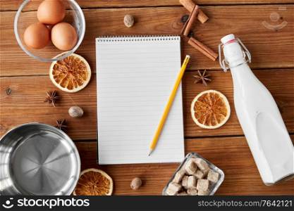 cooking, christmas and seasonal drinks concept - notebook with pencil, ingredients for eggnog, pot on wooden background. notebook, pencil, ingredients for eggnog cooking