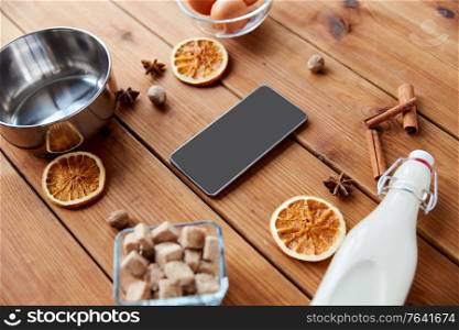 cooking, christmas and seasonal drinks concept - smartphone, ingredients for eggnog, pot on wooden background. smartphone, ingredients for eggnog cooking