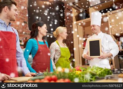 cooking class, culinary, food and people concept - happy friends and chef cook with blank menu chalk board in kitchen over snow effect