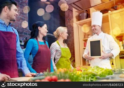 cooking class, culinary, food and people concept - happy friends and chef cook with blank menu chalk board in kitchen. happy friends and chef cook with menu in kitchen