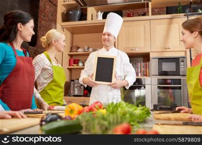 cooking class, culinary, food and people concept - happy women and chef cook with blank menu chalk board in kitchen