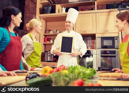 cooking class, culinary, food and people concept - happy women and chef cook with blank menu chalk board in kitchen