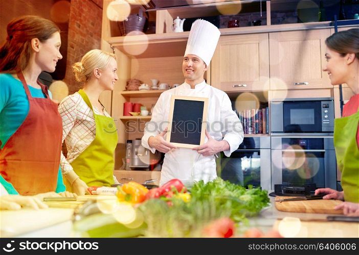 cooking class, culinary, food and people concept - happy women and chef cook with blank menu chalk board in kitchen. happy women and chef cook with menu in kitchen