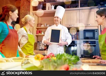 cooking class, culinary, food and people concept - happy women and chef cook with blank menu chalk board in kitchen. happy women and chef cook with menu in kitchen