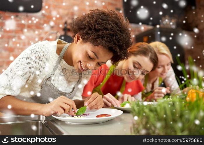 cooking class, friendship, food and people concept - happy women cooking and decorating plates with dishes in kitchen over snow effect
