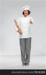 cooking, culinary and bakery concept - happy smiling female chef or baker in toque holding french bread or baguette and showing thumbs up over grey background. happy female chef with french bread or baguette