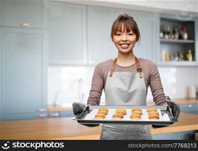 cooking, culinary and bakery concept - happy smiling female chef or baker in apron holding baking tray with oatmeal cookies over kitchen background. happy female in apron with cookies on oven tray