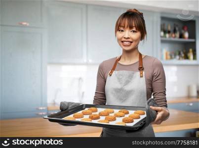 cooking, culinary and bakery concept - happy smiling woman or baker in apron holding baking tray with oatmeal cookies over home kitchen background. happy woman with cookies on oven tray at kitchen