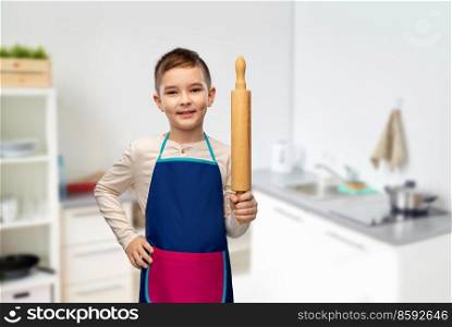 cooking, culinary and baking concept - happy smiling little boy in apron with rolling pin over home kitchen background. smiling boy in apron with rolling pin in kitchen